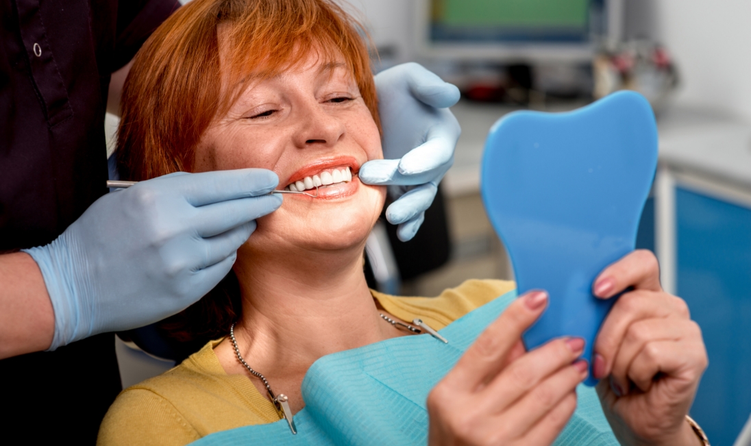 Older redheaded woman looking at her smile in mirror while sitting in dental chair