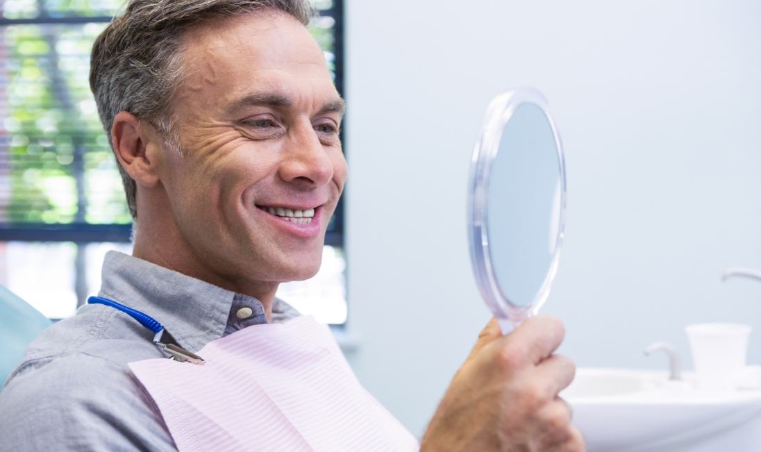 Older man in dental chair looking at his smile in a mirror